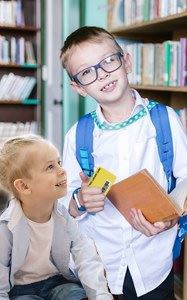 Two children in a library with a book and a library card, smiling.
